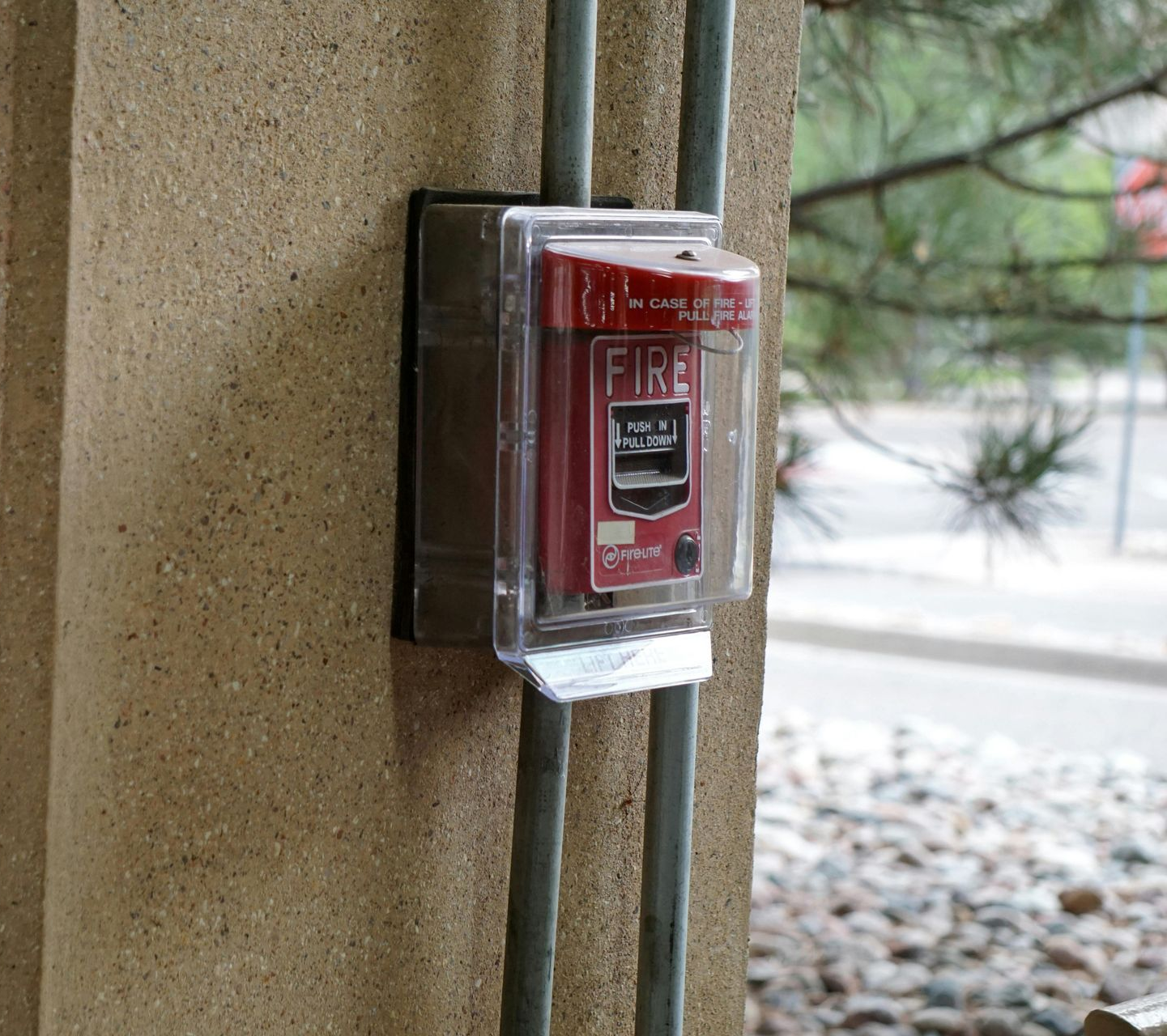 Red fire alarm pull station mounted on a wall by exposed conduit and a window.