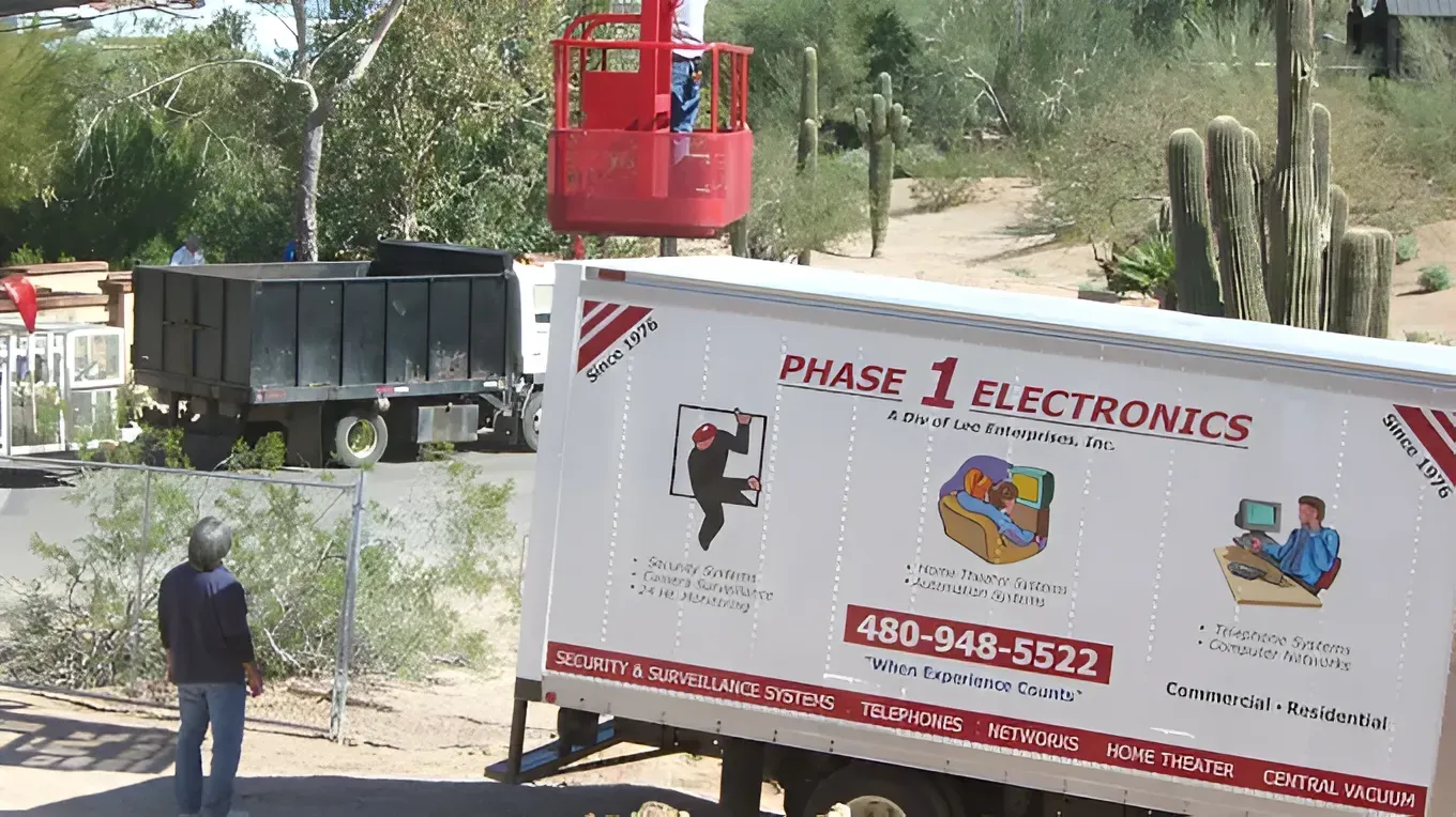 Construction site with a red lift beside a white Phase 1 Electronics truck and a black dump truck.