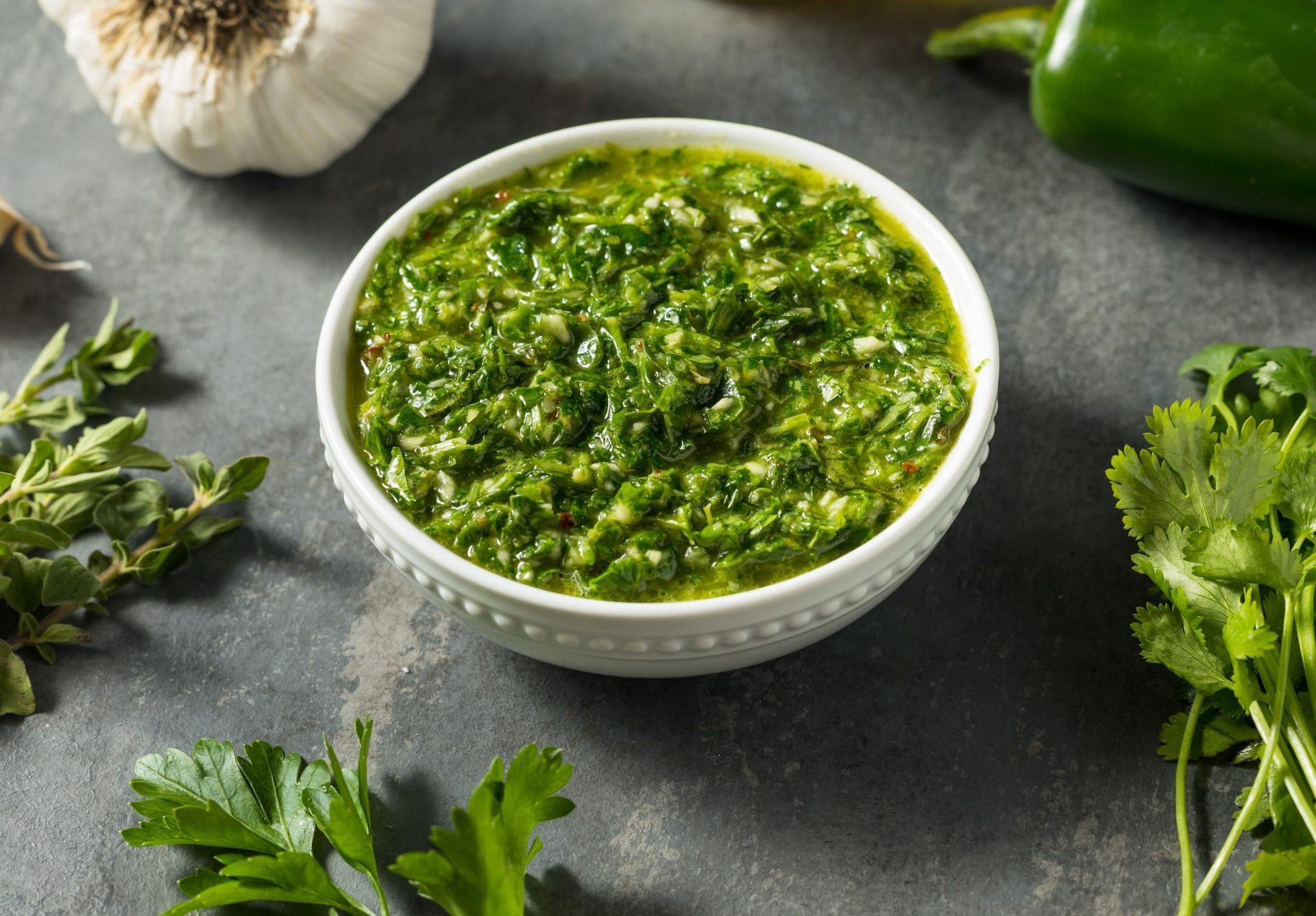 Bowl of chimichurri sauce with garlic, chili, and cilantro around it