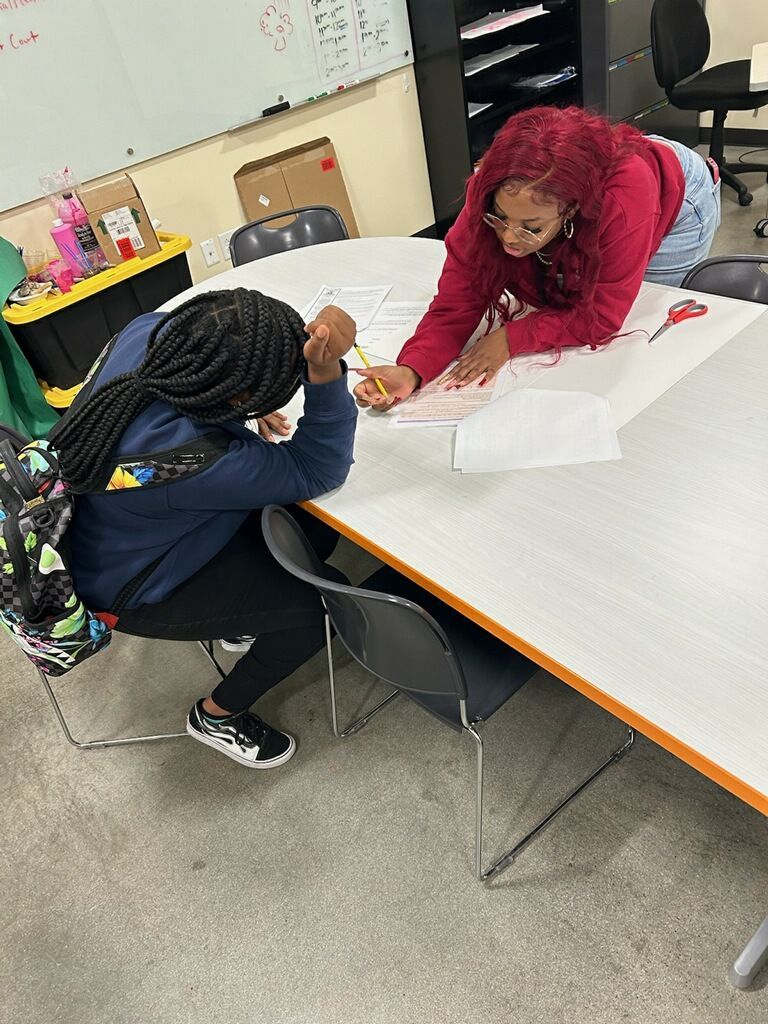 Two students leaning over a table, working together on a worksheet in a classroom.
