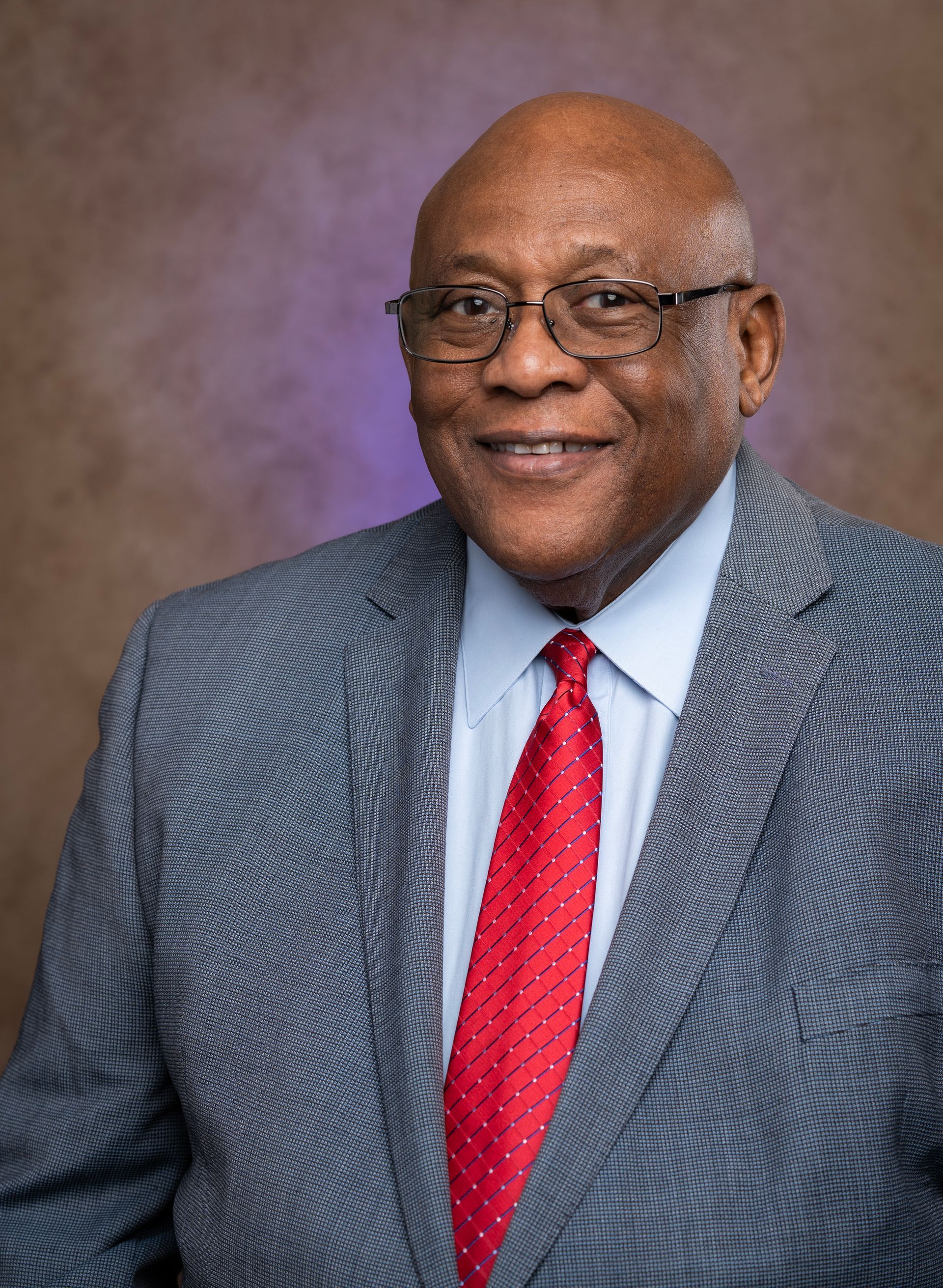 Smiling man in glasses, gray suit, white shirt, and red tie against a studio backdrop.