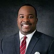 Portrait of a smiling man in a dark suit, white shirt, and red striped tie on a dark gray background