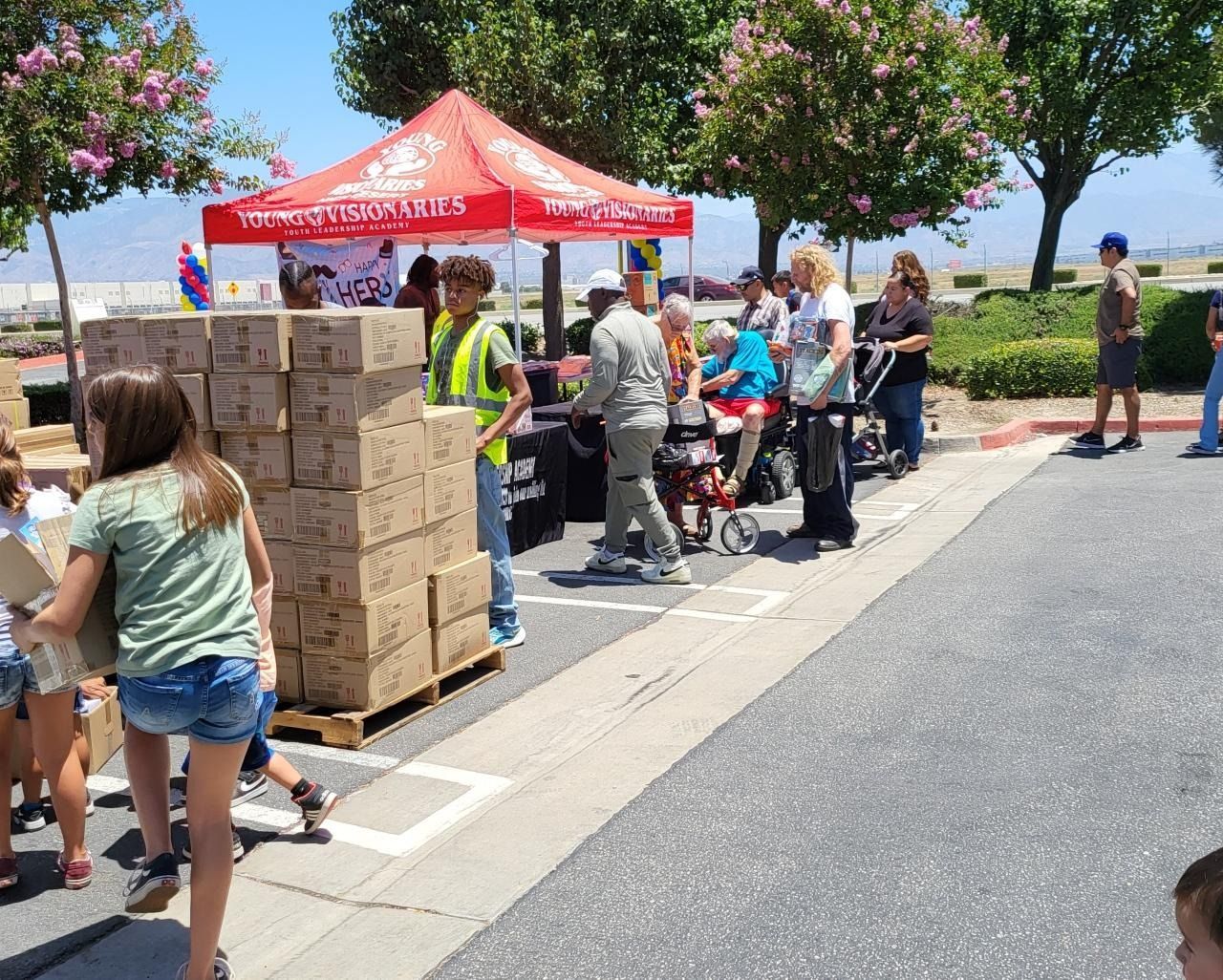 People gathered at a red canopy tent beside stacked boxes on a sunny street, with volunteers helping.