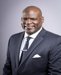 Studio portrait of a smiling man in a dark suit, white shirt, tie, and pocket square on a gray background