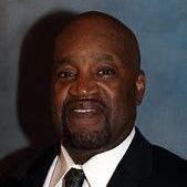 Portrait of a man in a dark suit, white shirt, and tie against a blue-gray background.