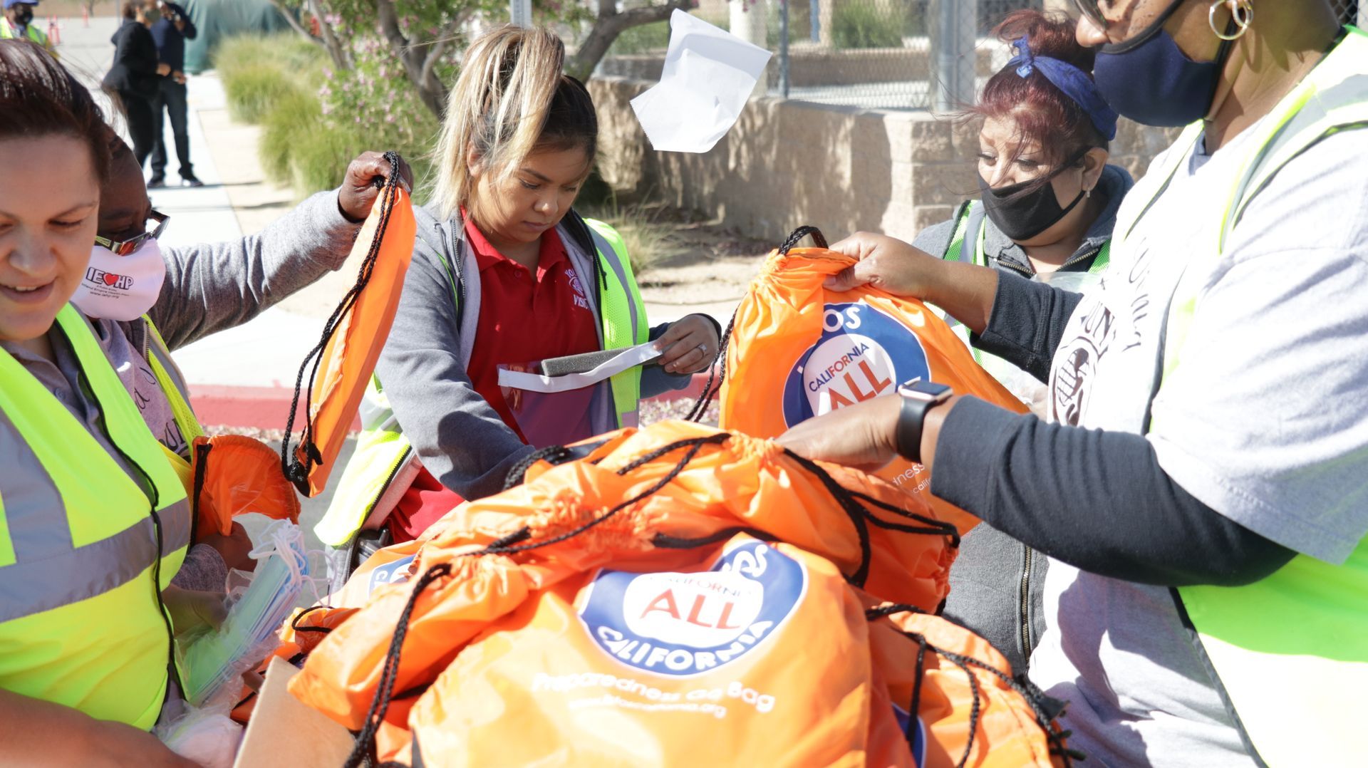 Volunteers in safety vests and masks load orange relief bags from a cart outdoors.