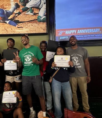 Group holding certificates poses indoors near a TV screen displaying “Happy Anniversary” and sports footage.