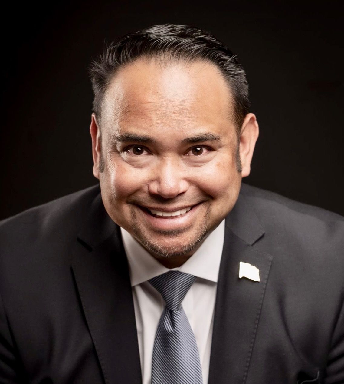 Smiling man in a dark suit and tie against a black background, with a small flag pin on his lapel
