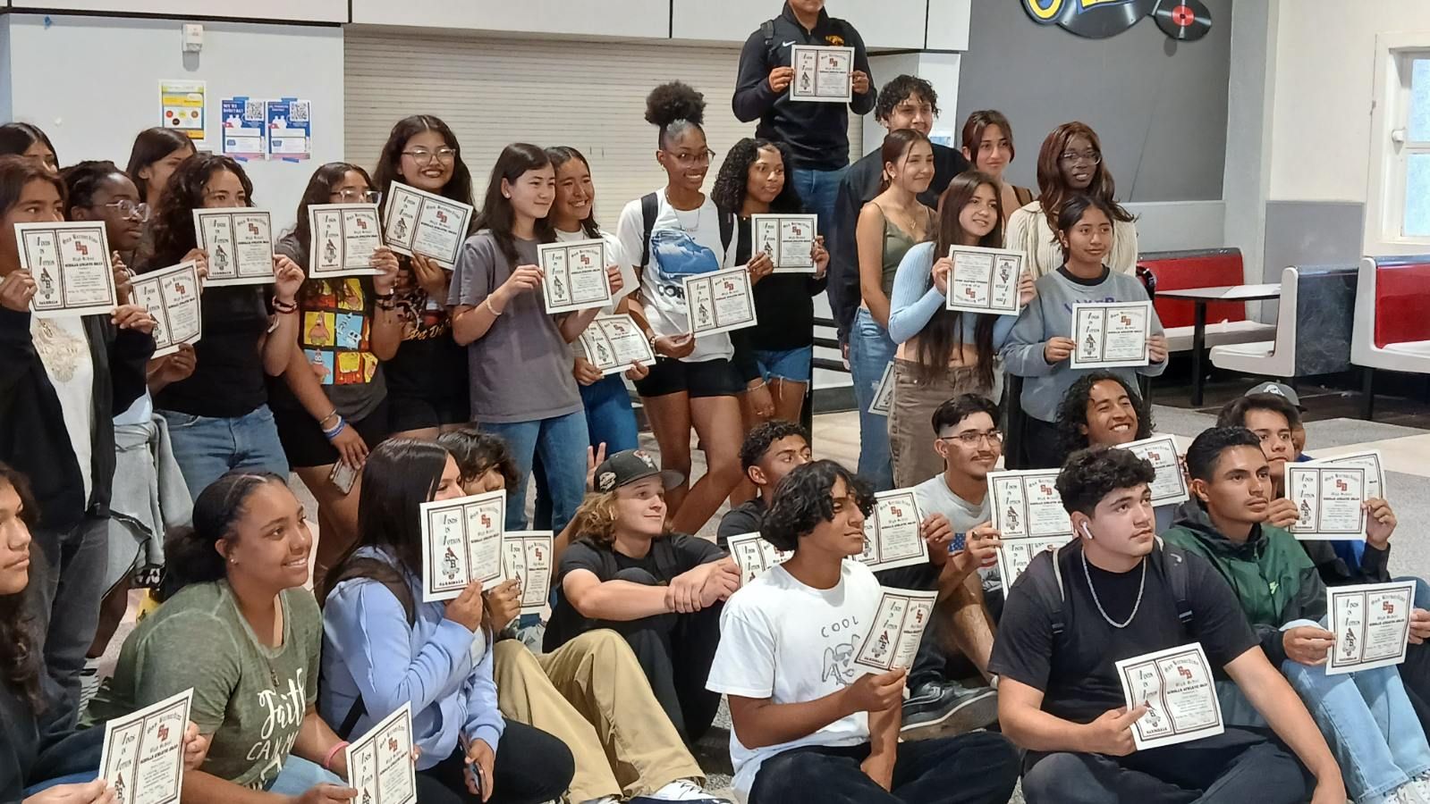 Group of people seated on bleachers, smiling and holding up small signs in a classroom or gym setting