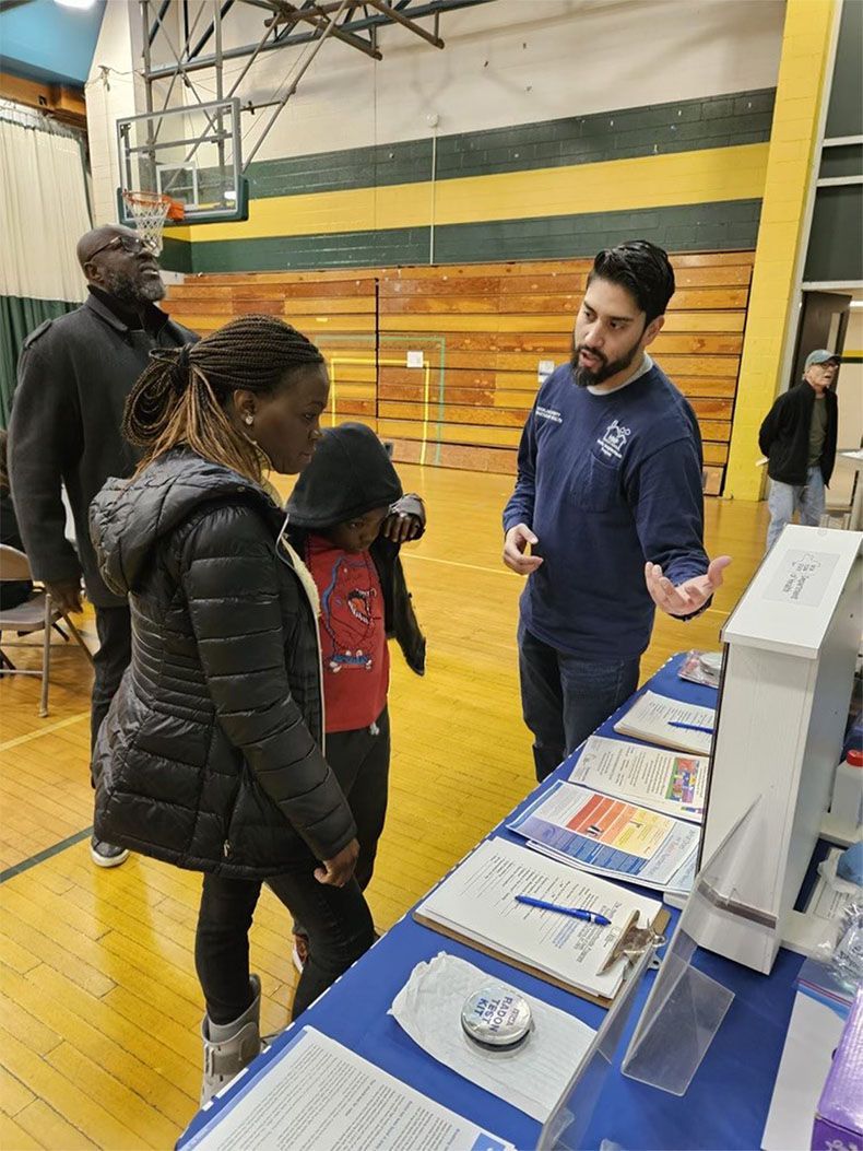 People at a school fair table with brochures, talking to a staff member in a gymnasium.