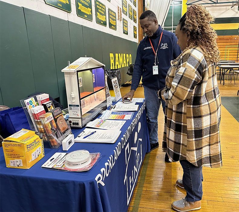 Two people at a school table display booth with brochures, flyers, and a monitor in a gymnasium.