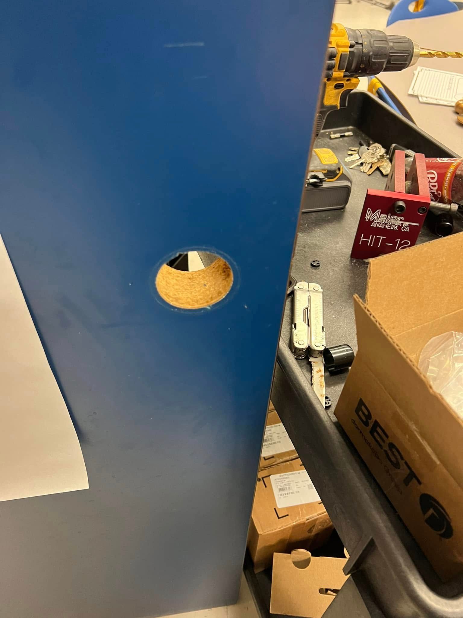 Blue partition with a round hole, beside a cluttered workshop table with tools and a cardboard box.
