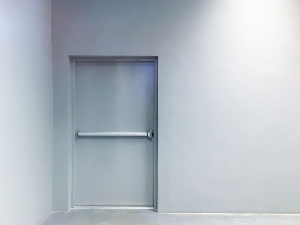 Closed gray door with a horizontal push bar in a plain white hallway