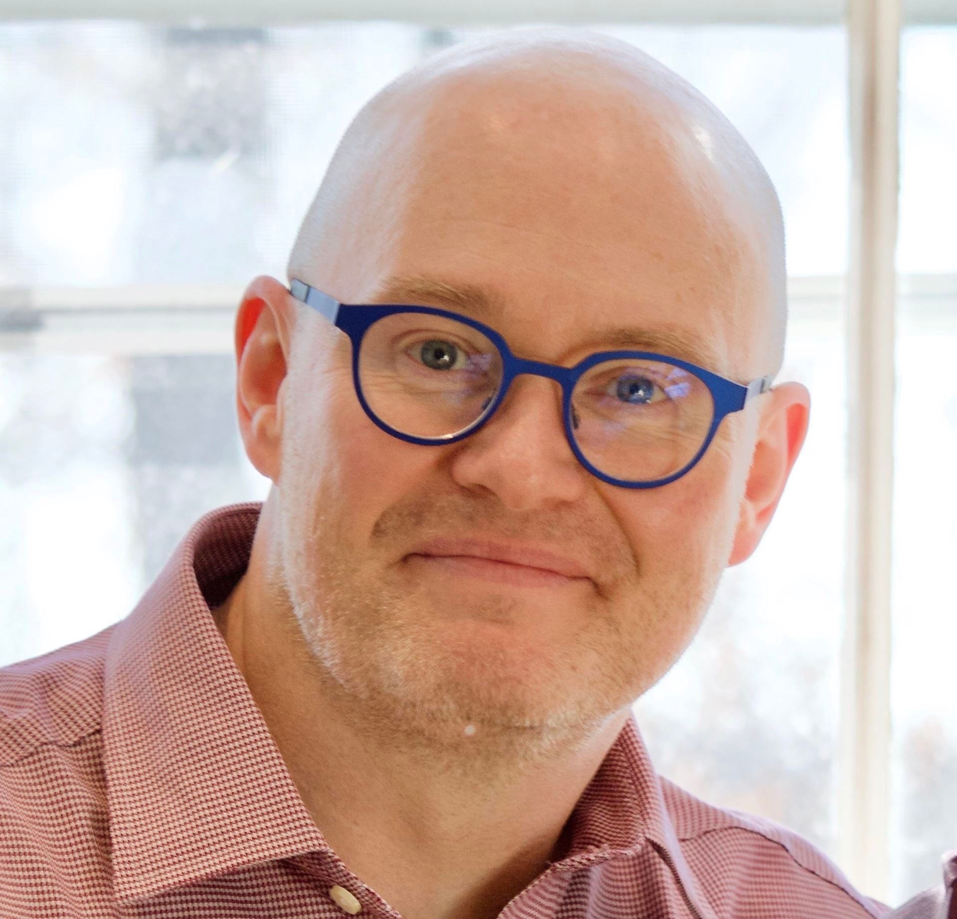 Close-up of a bald person wearing blue glasses and a red checkered shirt, smiling indoors near a window