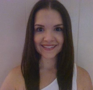 Smiling woman with long dark hair standing against a plain light wall, wearing a white top