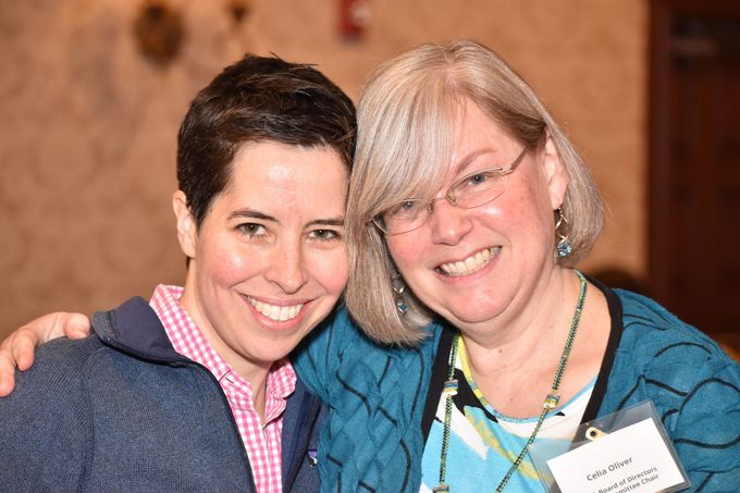 Two smiling people posing together at an indoor event, one in a navy sweater and one in a teal cardigan.