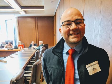 Man in glasses and red tie stands in a conference room, smiling at the camera.
