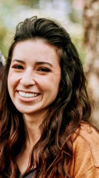 Smiling person with long dark hair wearing a brown top outdoors