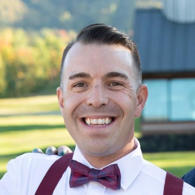 Smiling man in a white shirt and burgundy bow tie outdoors, with a blurred building in the background