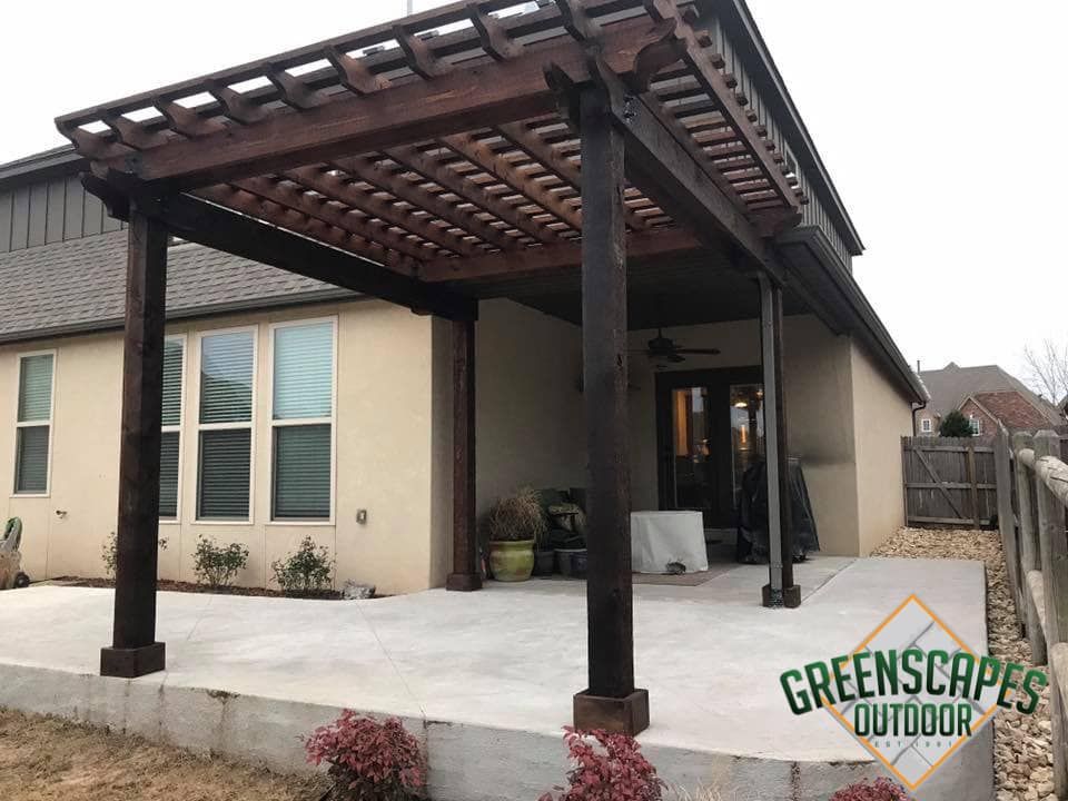 Dark stained wooden pergola over a concrete patio; attached to beige house.