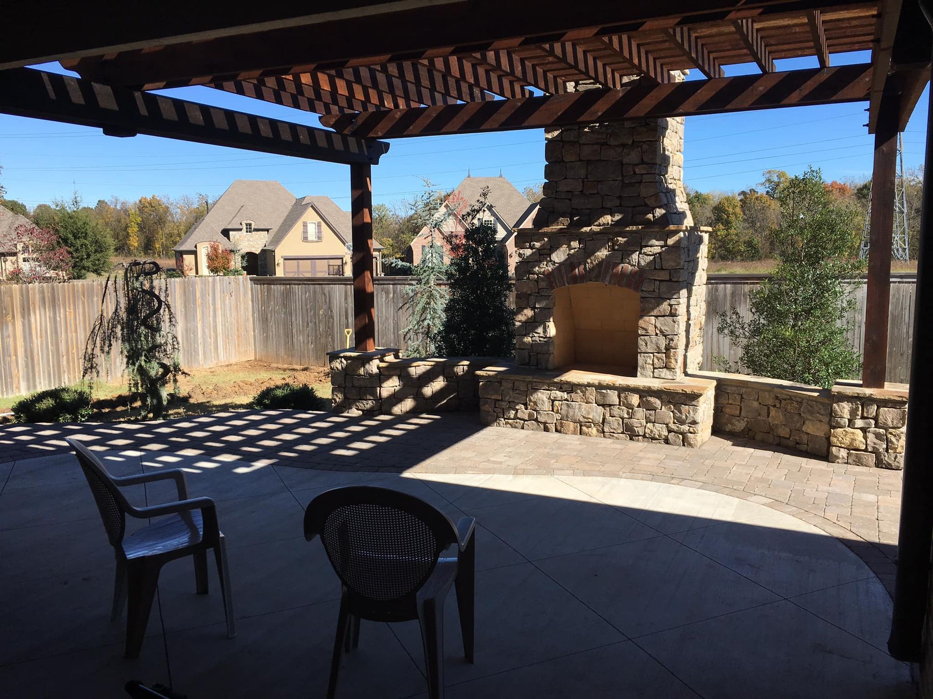 Outdoor patio with pergola, stone fireplace, and two chairs. Houses and trees visible in background.