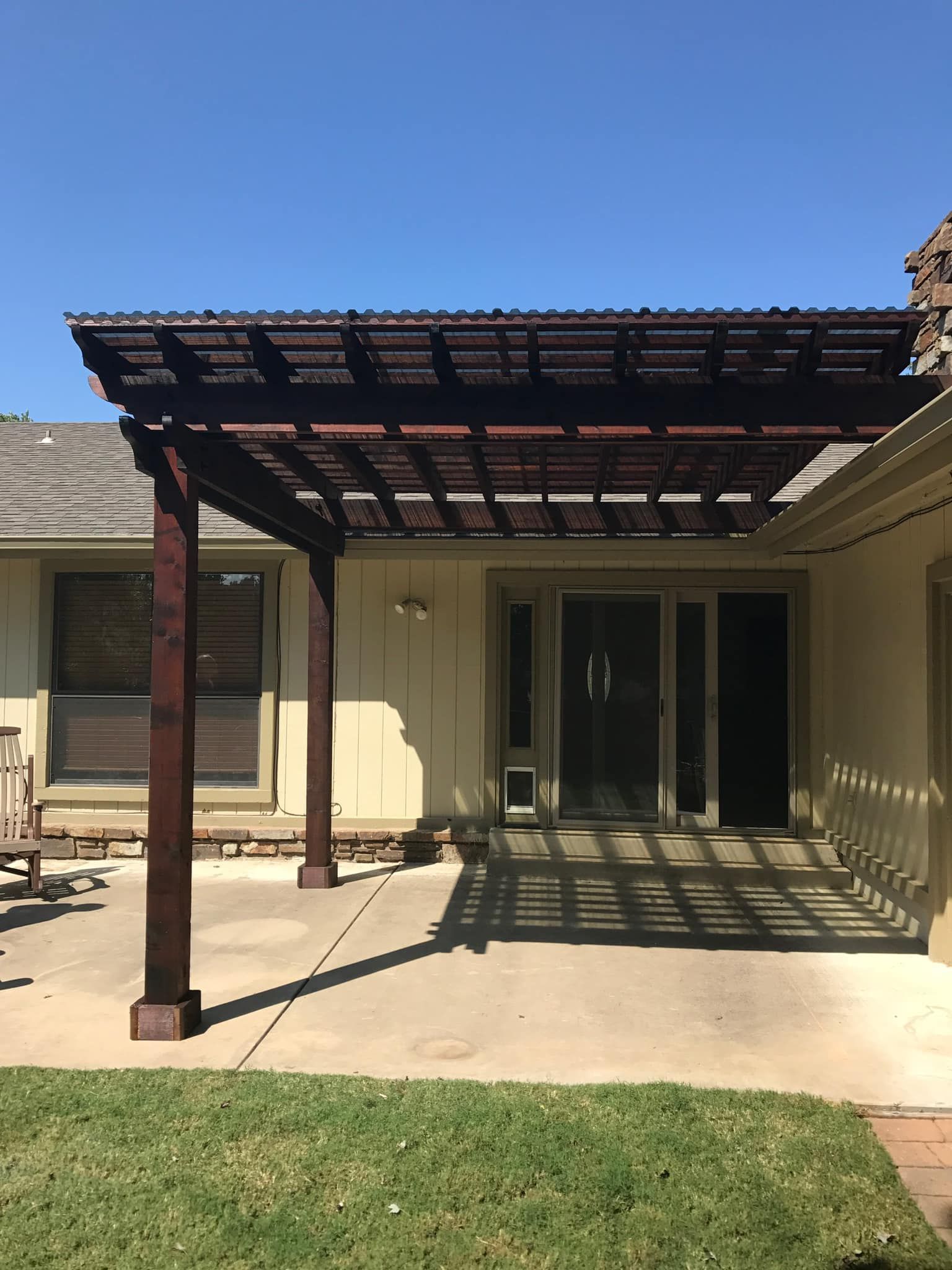 Pergola over patio, casting shadow on concrete. Dark wood structure, tan house, blue sky, green grass.