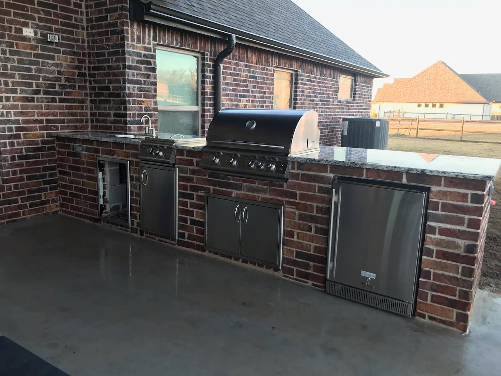 A brick outdoor kitchen with a grill and a refrigerator.