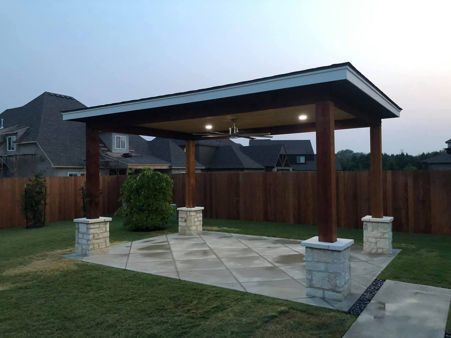 Patio with a dark wooden frame and stone-covered pillars, under a roof, and concrete floor on a green lawn.
