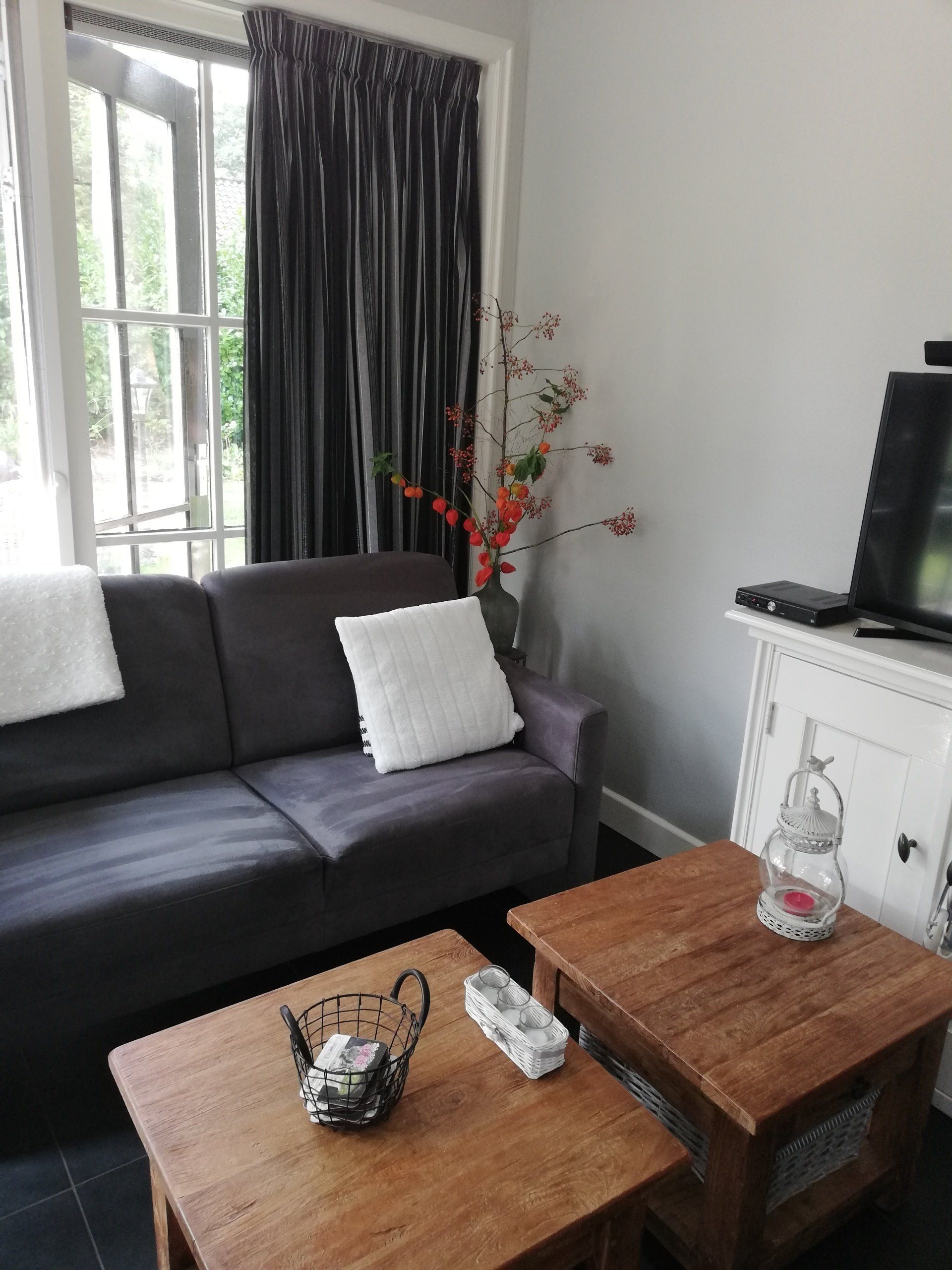 Woonkamer met grijze bank, kleine houten tafeltjes, zwarte gordijnen en een televisie.