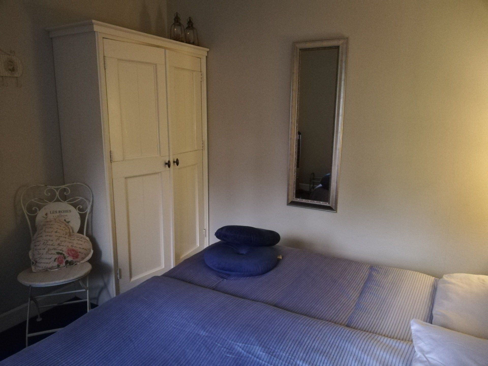 Slaapkamer met een witte kledingkast, een bed met blauw beddengoed, een spiegel en een decoratieve stoel.