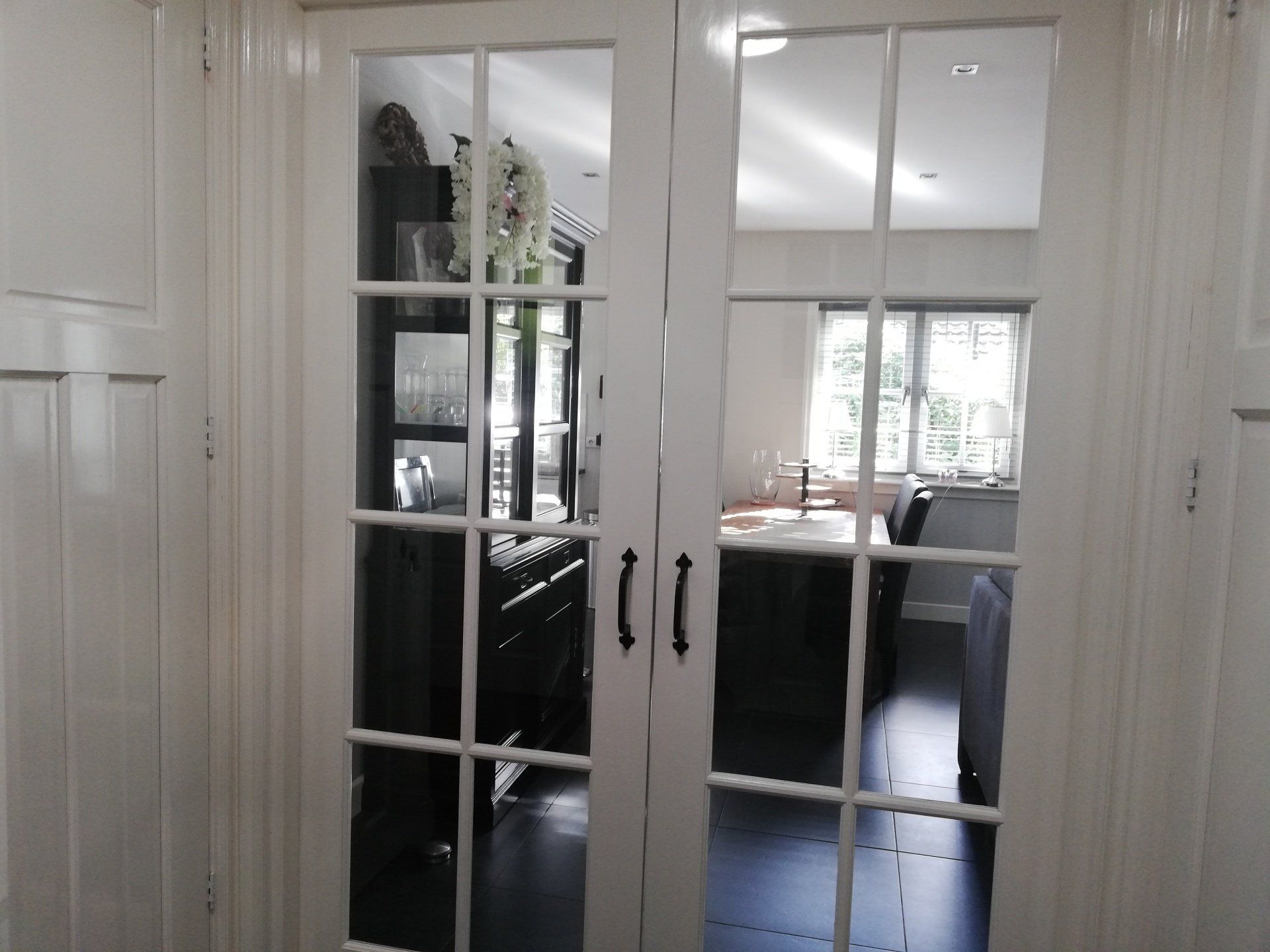 Witte openslaande deuren met meerdere ruiten, die een kamer met donker meubilair en een keukenhoek weerspiegelen.