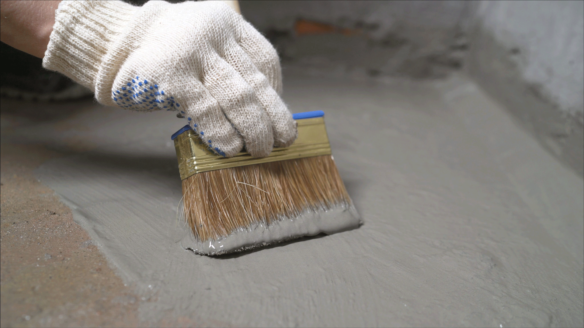 Hand in glove with brush spreading gray cement on a concrete surface.
