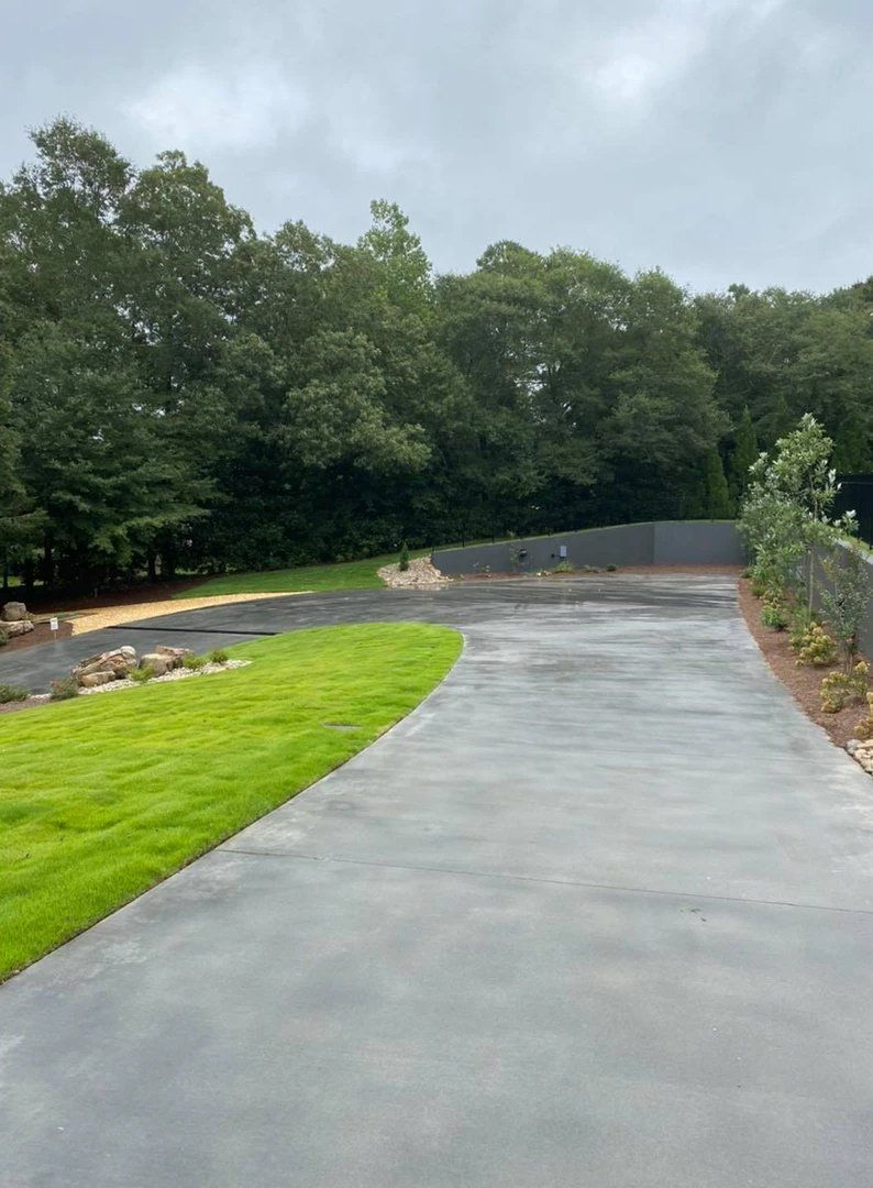 A concrete driveway leading to a lush green field surrounded by trees on a cloudy day.