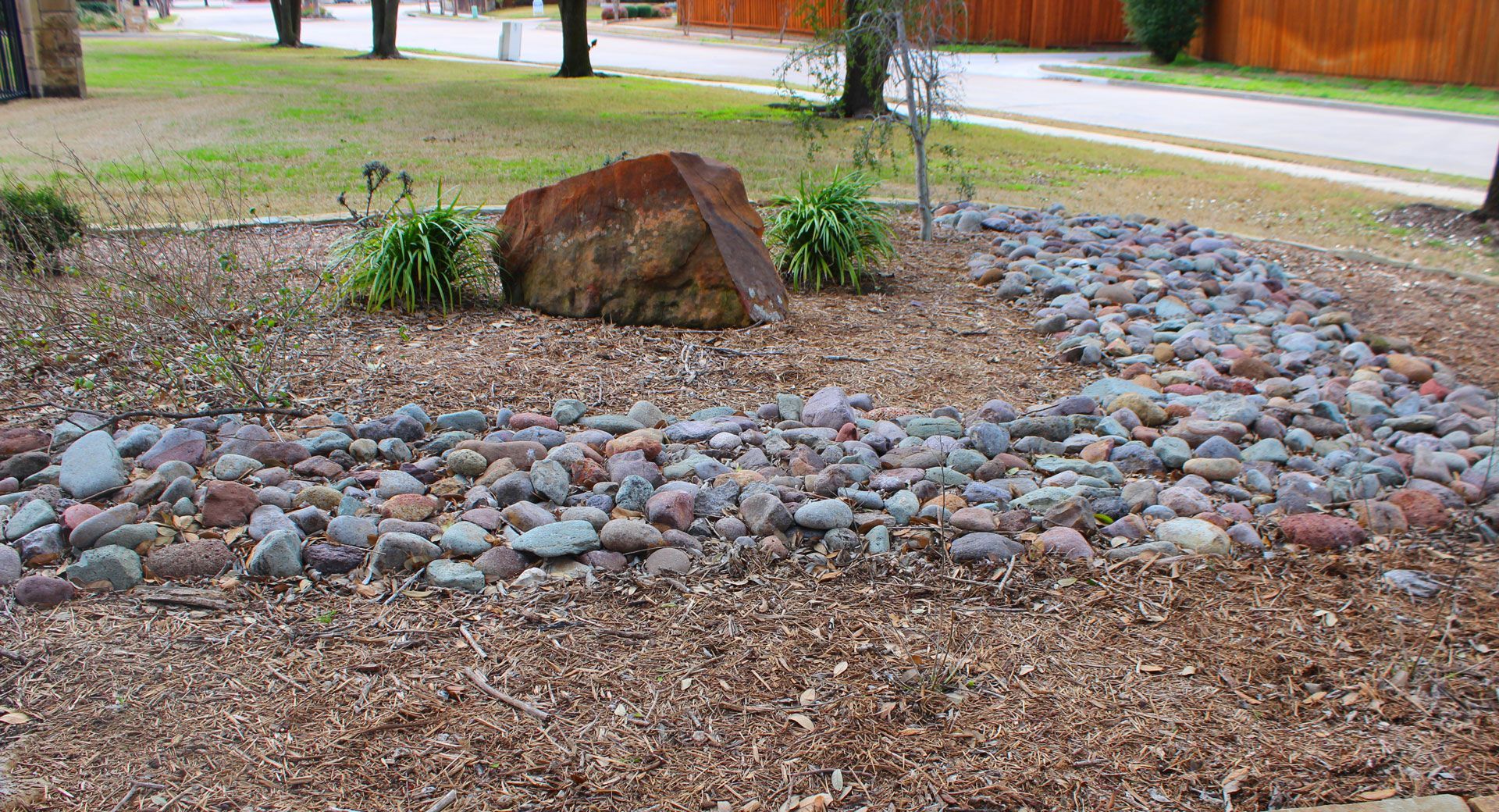 Rock garden with large boulder, small plants, and a curved border of gray stones in a yard.
