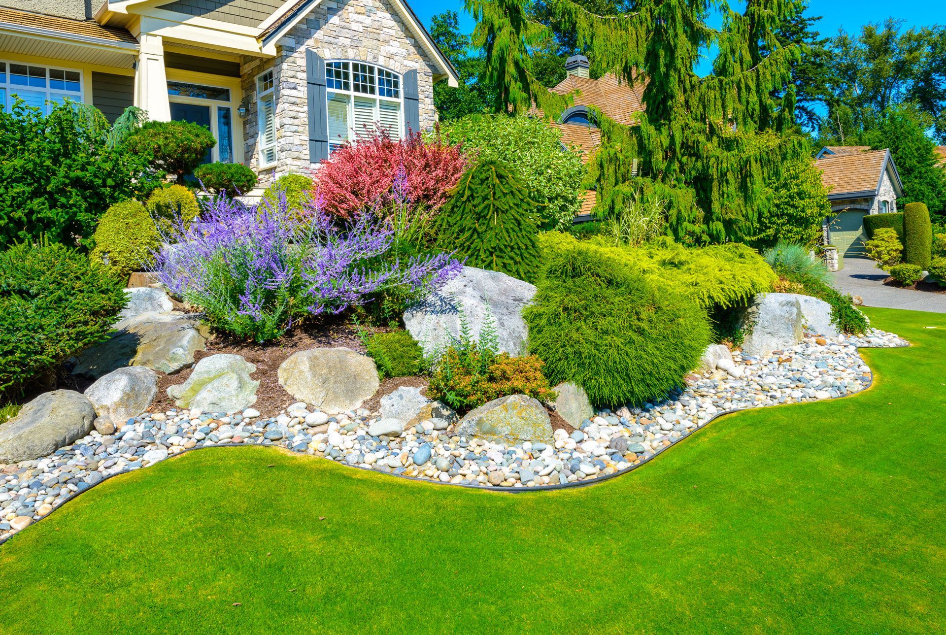 Colorful landscaped front yard with flowering shrubs, rocks, and a house in the background