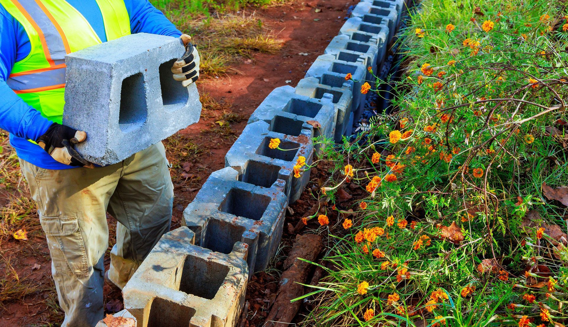 Worker carrying cinder blocks beside a garden bed with flowers and a stacked block wall