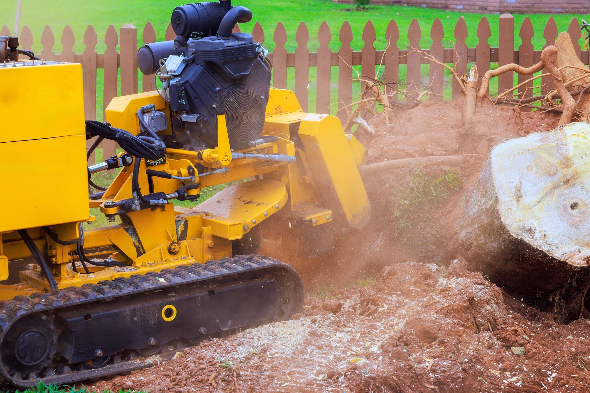Yellow bulldozer clearing dirt beside a large rock, kicking up dust near a wooden fence.
