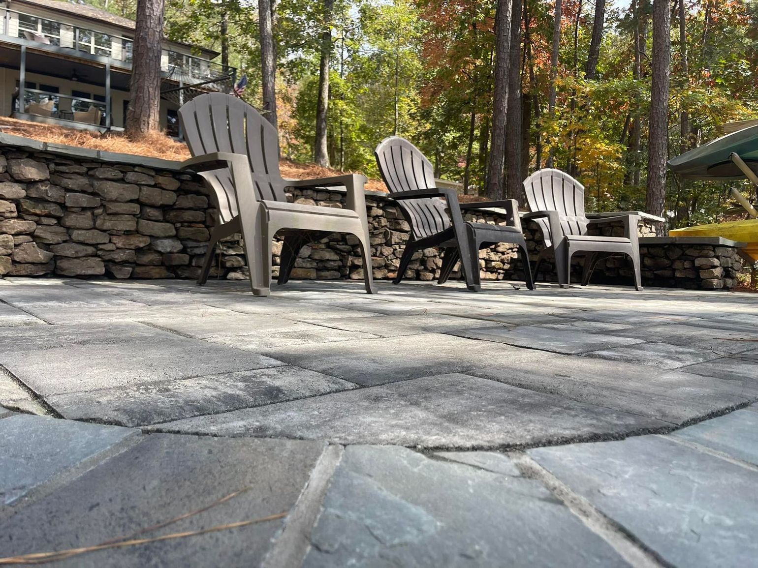 Patio with three white Adirondack chairs on stone pavers beside a retaining wall and trees