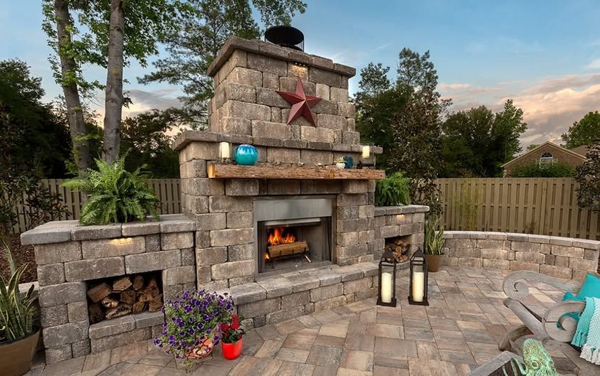 Stone outdoor fireplace with flames, lanterns, and potted flowers on a patio at sunset