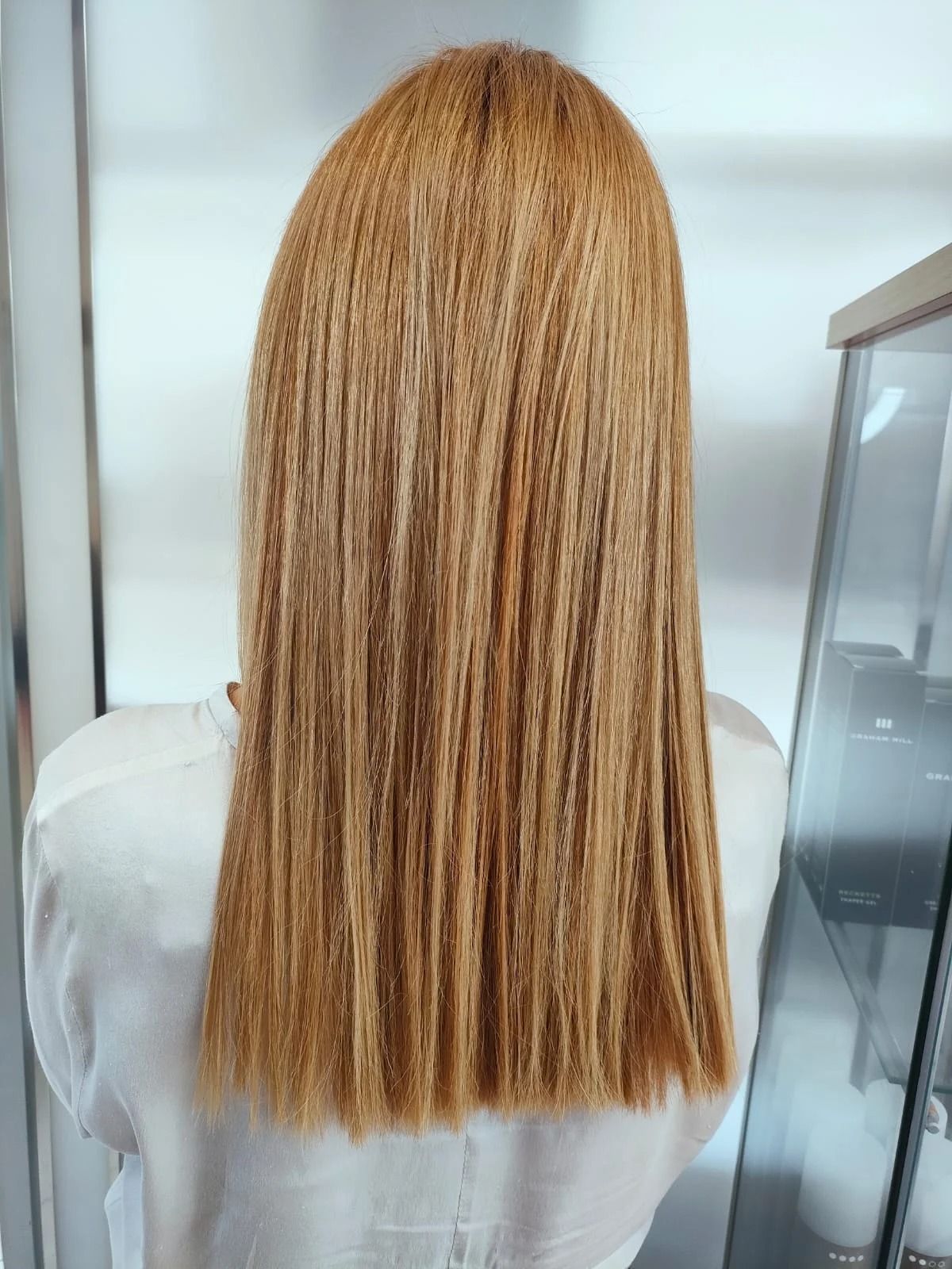 Donna con capelli lisci, castano dorato. Vista posteriore, con indosso una camicia bianca, all'interno di un salone di bellezza.