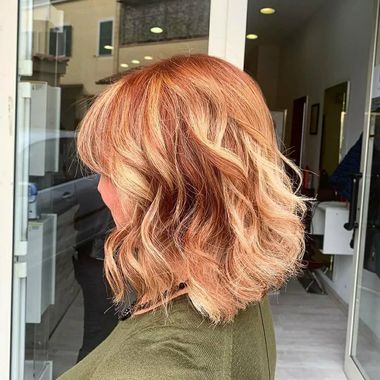 Donna con capelli biondo-rossicci ondulati, lunghi fino alle spalle, davanti alla vetrina di un salone.