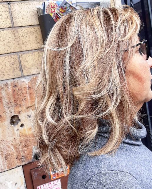 Donna con capelli biondi e castani ondulati e scalati, occhiali da sole e dolcevita grigio. Sfondo muro di mattoni.
