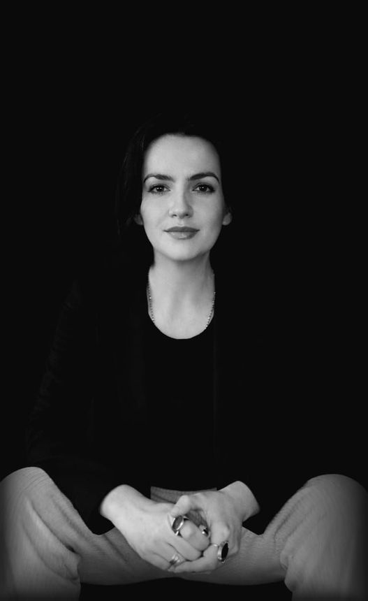 Woman seated, wearing a black shirt and light-colored pants, hands clasped, looking at the camera.