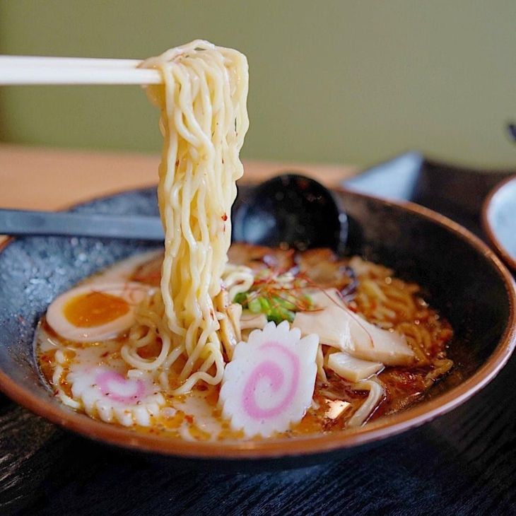 Chopsticks lifting noodles from a bowl of ramen with egg, fish cake, and broth