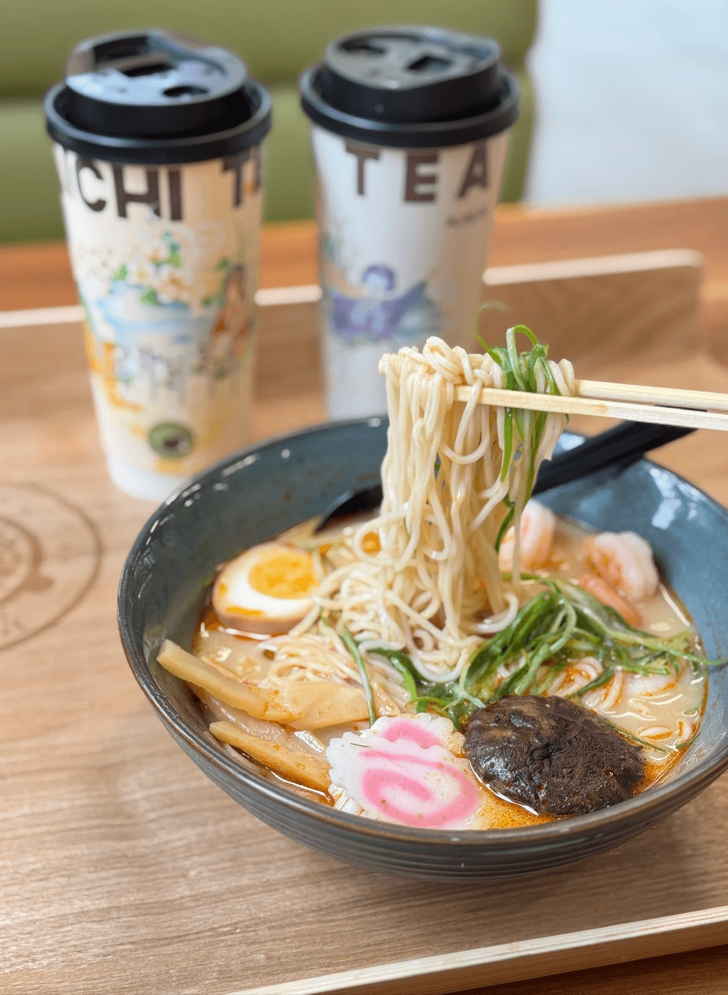 Chopsticks lifting ramen from a black bowl beside tea cups on a wooden table