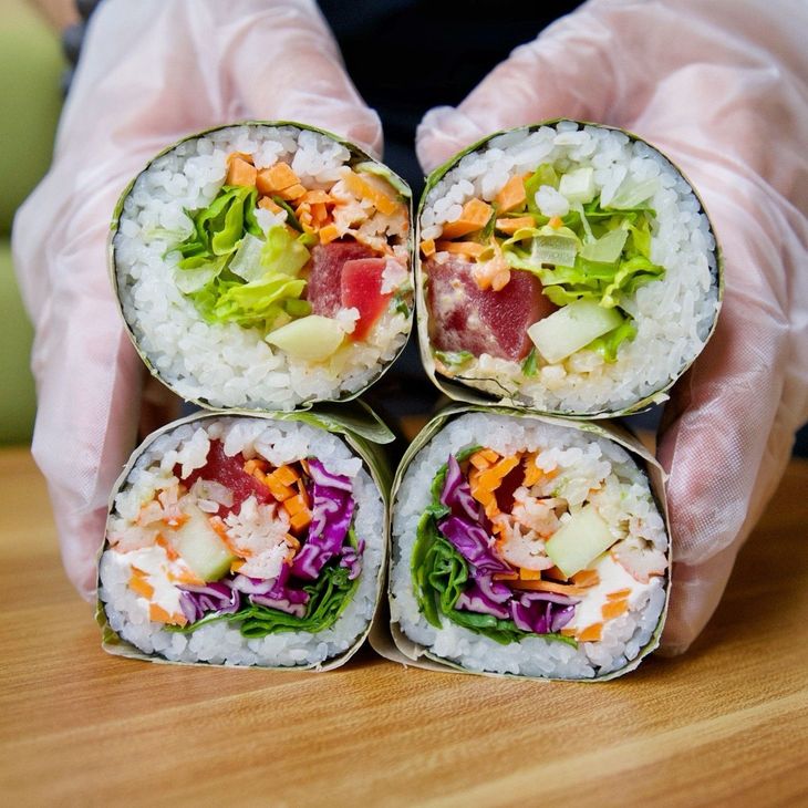 Gloved hands holding four cut sushi rolls with colorful vegetable filling on a wooden board