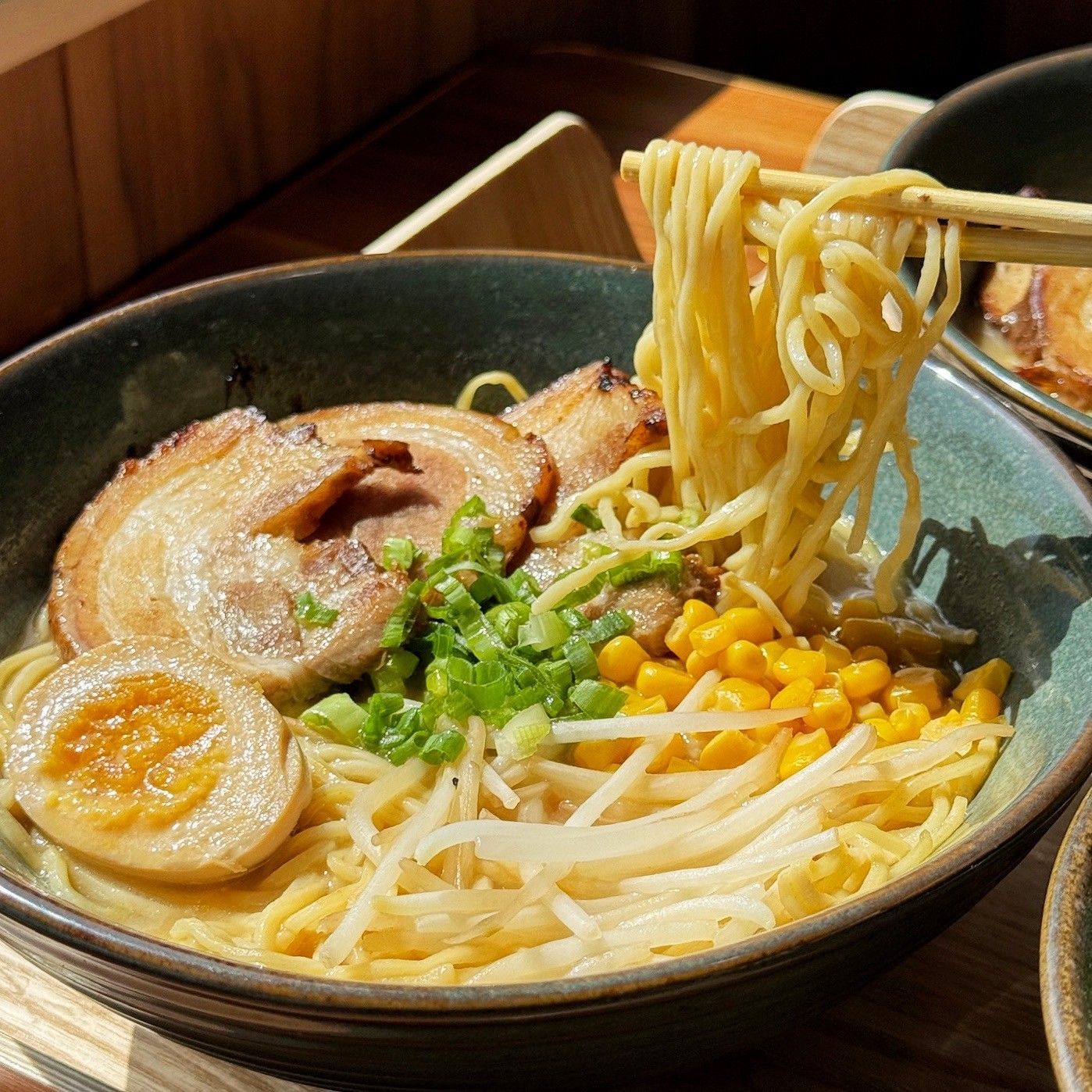 Bowl of ramen with sliced pork, egg, corn, bean sprouts, and green onions, with chopsticks lifting noodles