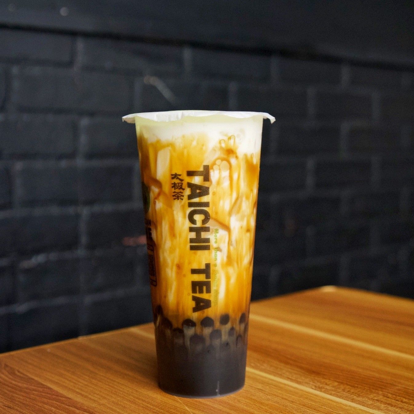 Tall bubble tea with caramel swirls and black tapioca pearls on a wooden table against a dark brick wall.