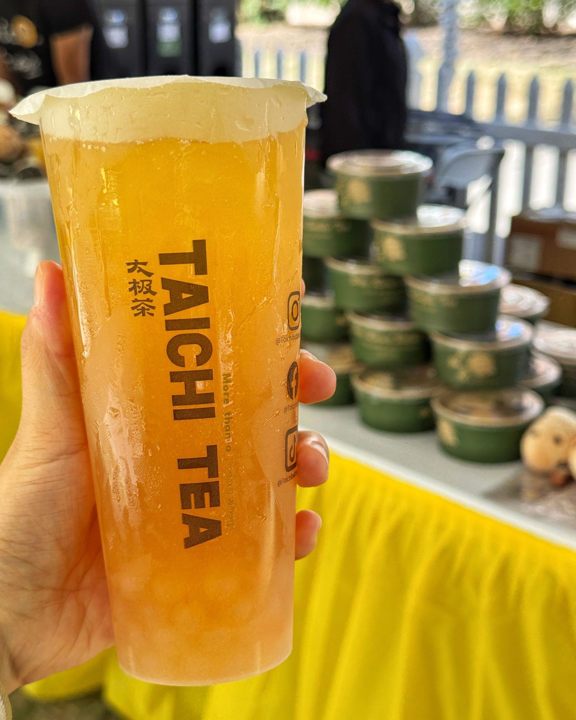 Hand holding a tall iced tea with foam, labeled “TAICHI TEA,” at an outdoor stall.