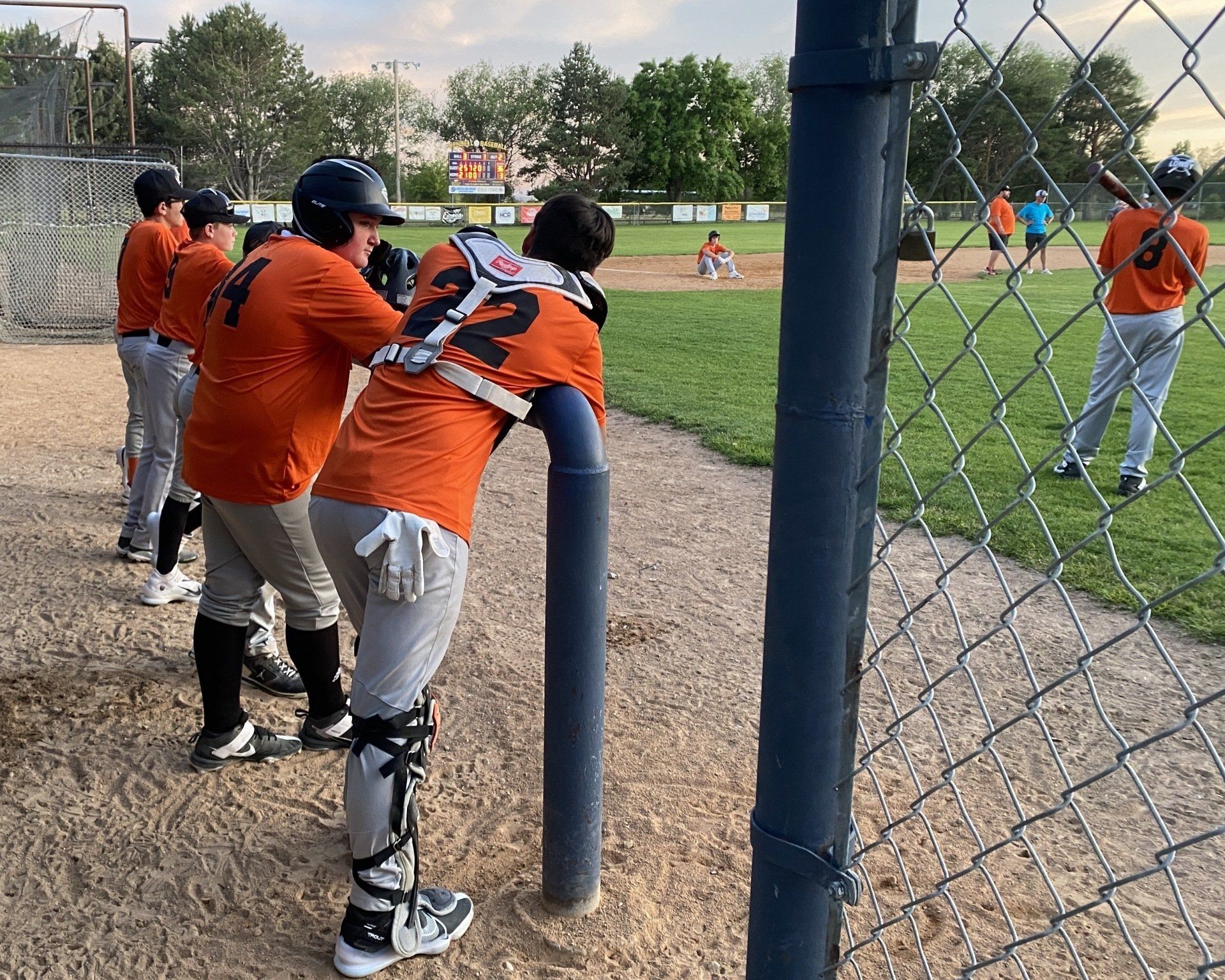 Baseball players in orange uniforms watch a game from the dugout, behind a fence.